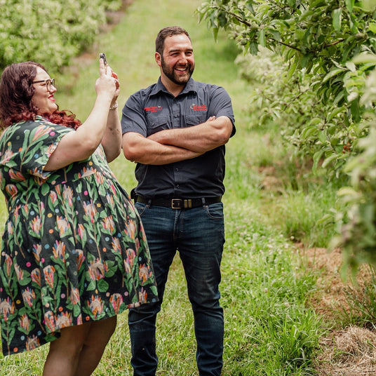 Two people standing in an orchard, one taking a photo with a smartphone.
