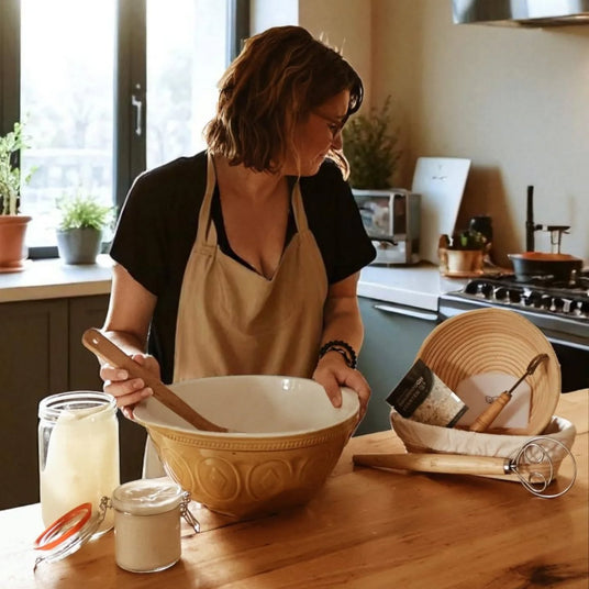 A woman wearing a beige apron stands in a warm kitchen, mixing ingredients in a large beige ceramic bowl with a wooden spoon. A matching woven basket and glass jars sit on the wooden counter beside her.