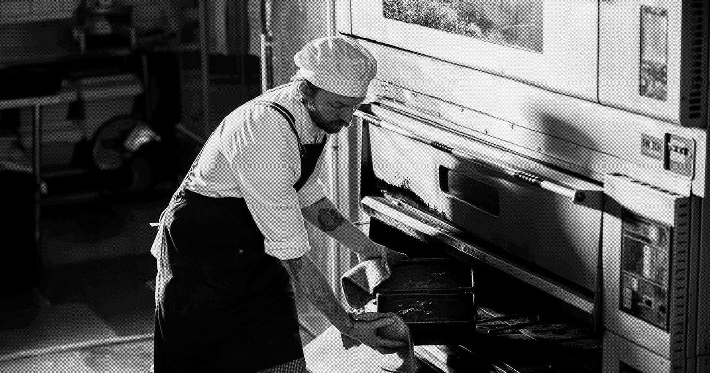 Person wearing a chef's hat and apron, reaching into an oven.