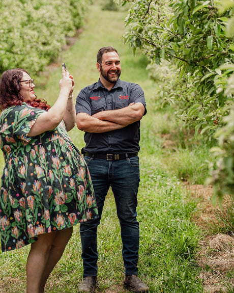 Two people standing in an orchard, one taking a photo with a smartphone.