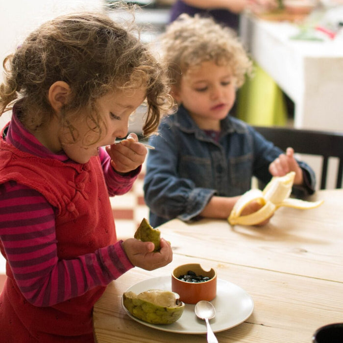Two children at a table with food, including a banana and a fruit bowl.