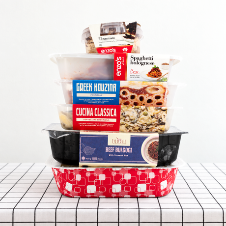 A stack of packaged meals from Dinner King, Foodee, Cucina Classica, Greek Kouzina and Enzo's at Home on a table