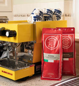 Yellow espresso machine with two red coffee bags labeled 'Cucina' on a kitchen counter.