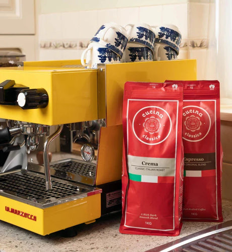Yellow espresso machine with two red coffee bags labeled 'Cucina' on a kitchen counter.