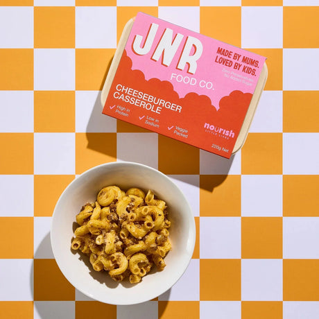 JNR food co cheeseburger casserole package next to a bowl of macaroni and cheese on a checkered background