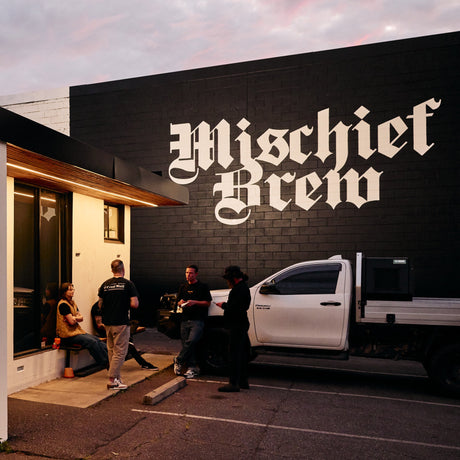 A black brick building with large white gothic lettering reading 'Mischief Brew' on the exterior wall. Several people stand and sit near a white pickup truck parked in front of the building during dusk.